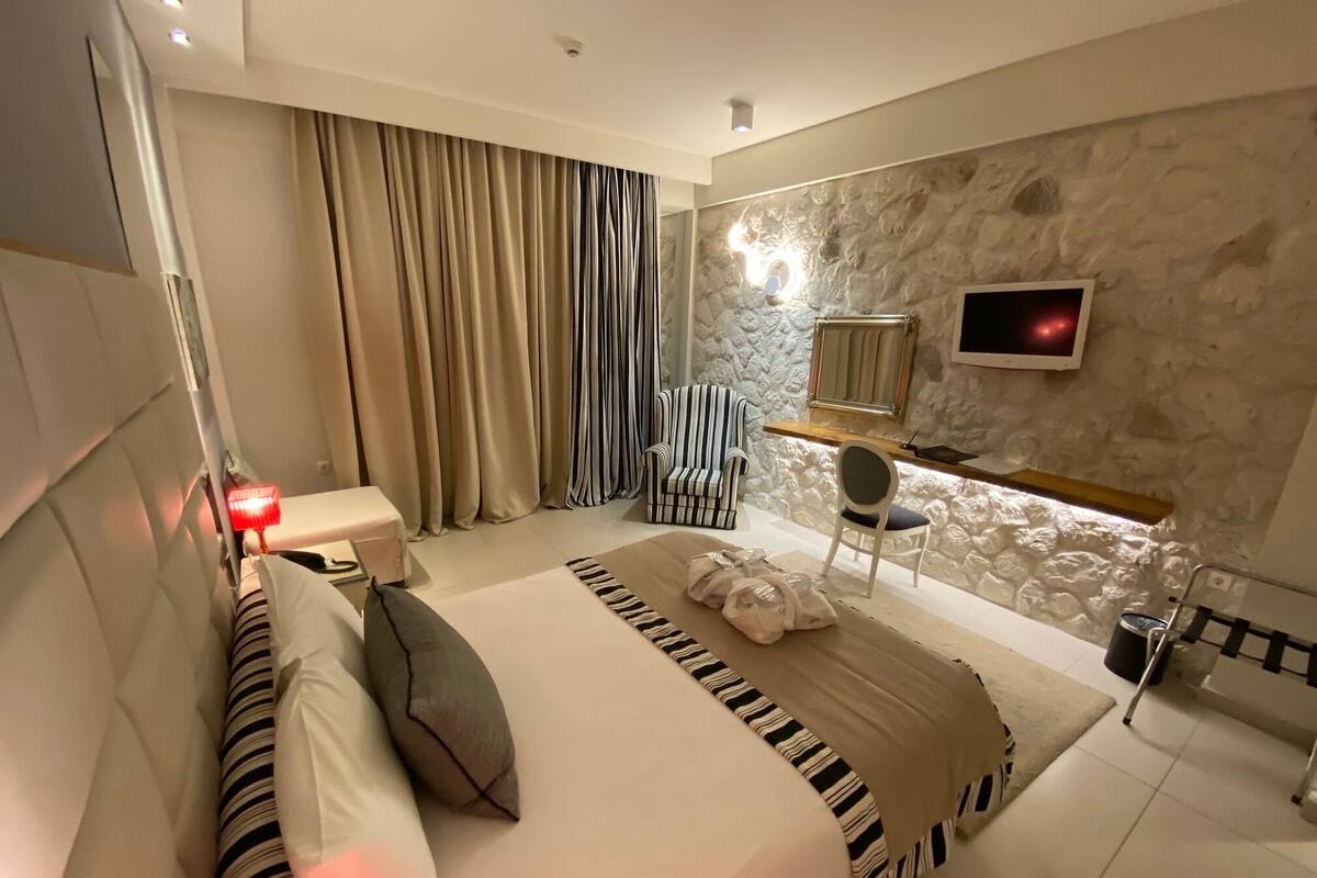 Ein modernes Hotelzimmer mit einem großen Bett, einem Nachttisch, einem Sessel, einem an einer Steinwand montierten Fernseher und einem Fenster mit Vorhängen.