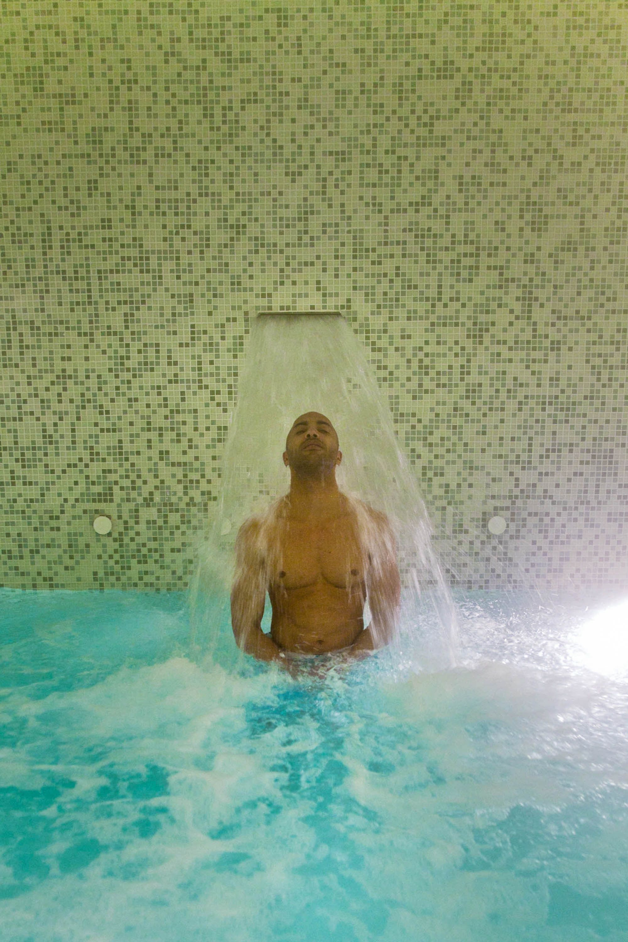 Una persona en una bañera con ducha sobre la cabeza, rodeada de un muro con mosaicos.
