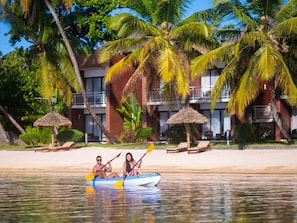 Una pareja en kayak en una playa tranquila con palmeras y un hotel al fondo.