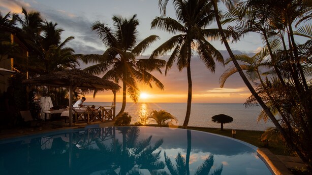 A poolside area with a sunset view, palm trees, and a thatched-roof structure.