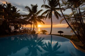 A poolside area with a sunset view, palm trees, and a thatched-roof structure. - Mantis Soanambo Hotel and Spa (Nosy Boraha)