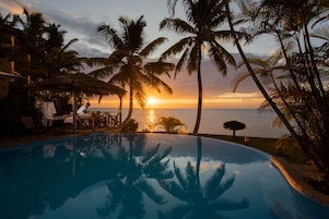 Zona de piscina con vista al atardecer, palmeras y una estructura de techo de paja.