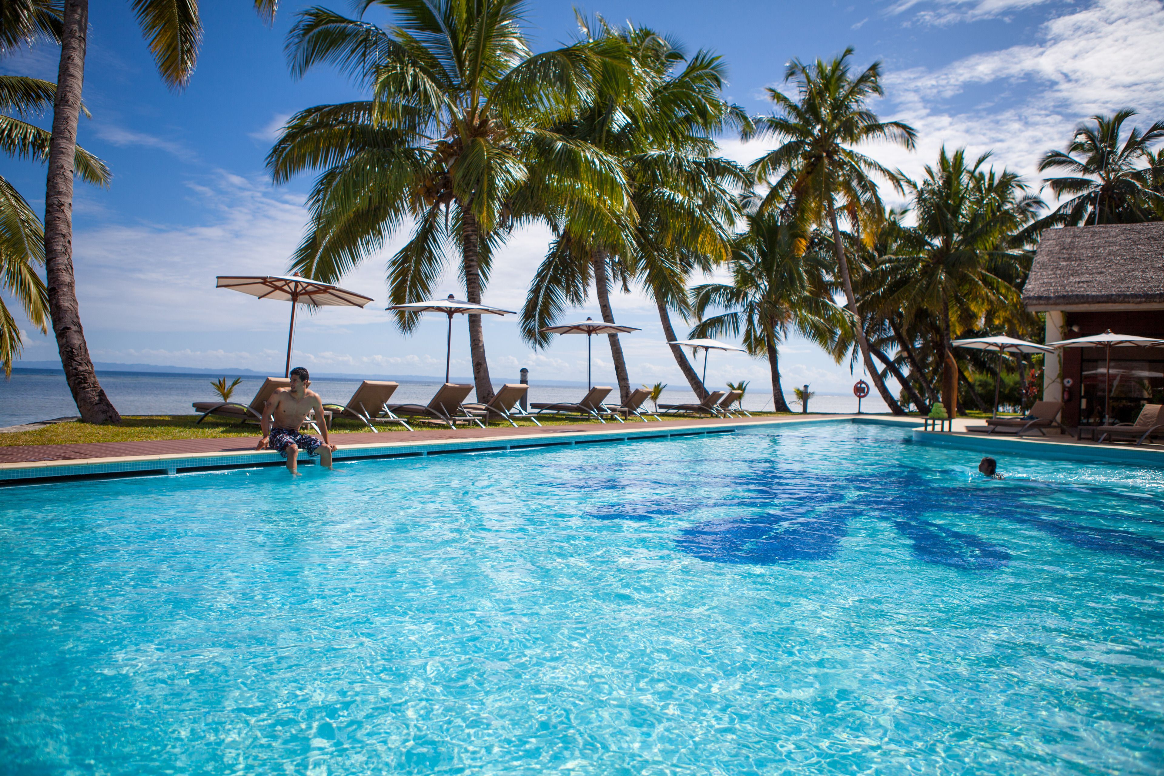 Una piscina con sillas de descanso y sombrillas, palmeras y vista al océano.