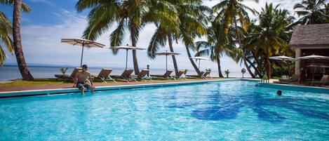 Una piscina con sillas de descanso y sombrillas, palmeras y vista al océano.