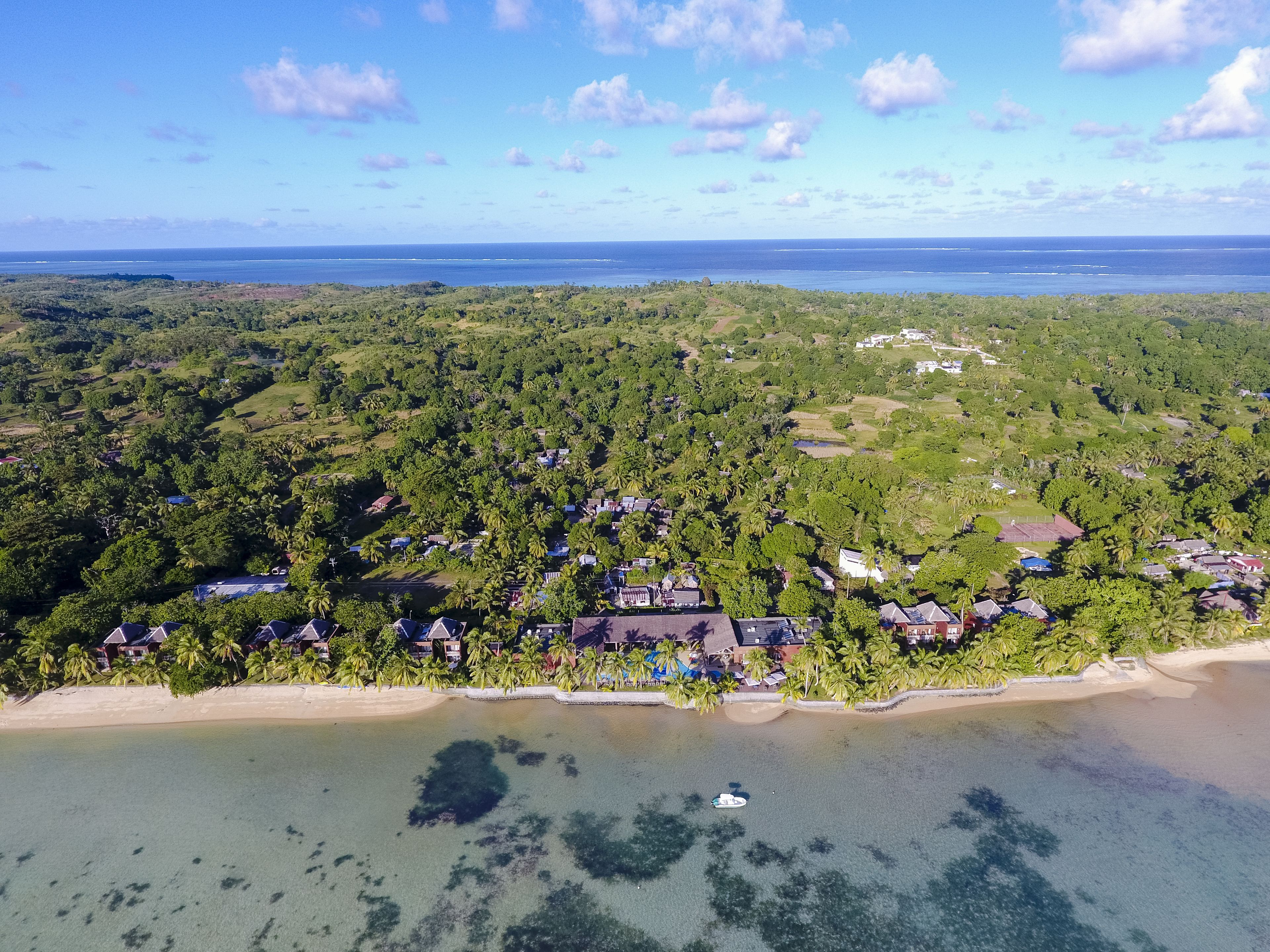An aerial view of a coastal area with dense greenery, a sandy beach, and scattered buildings.