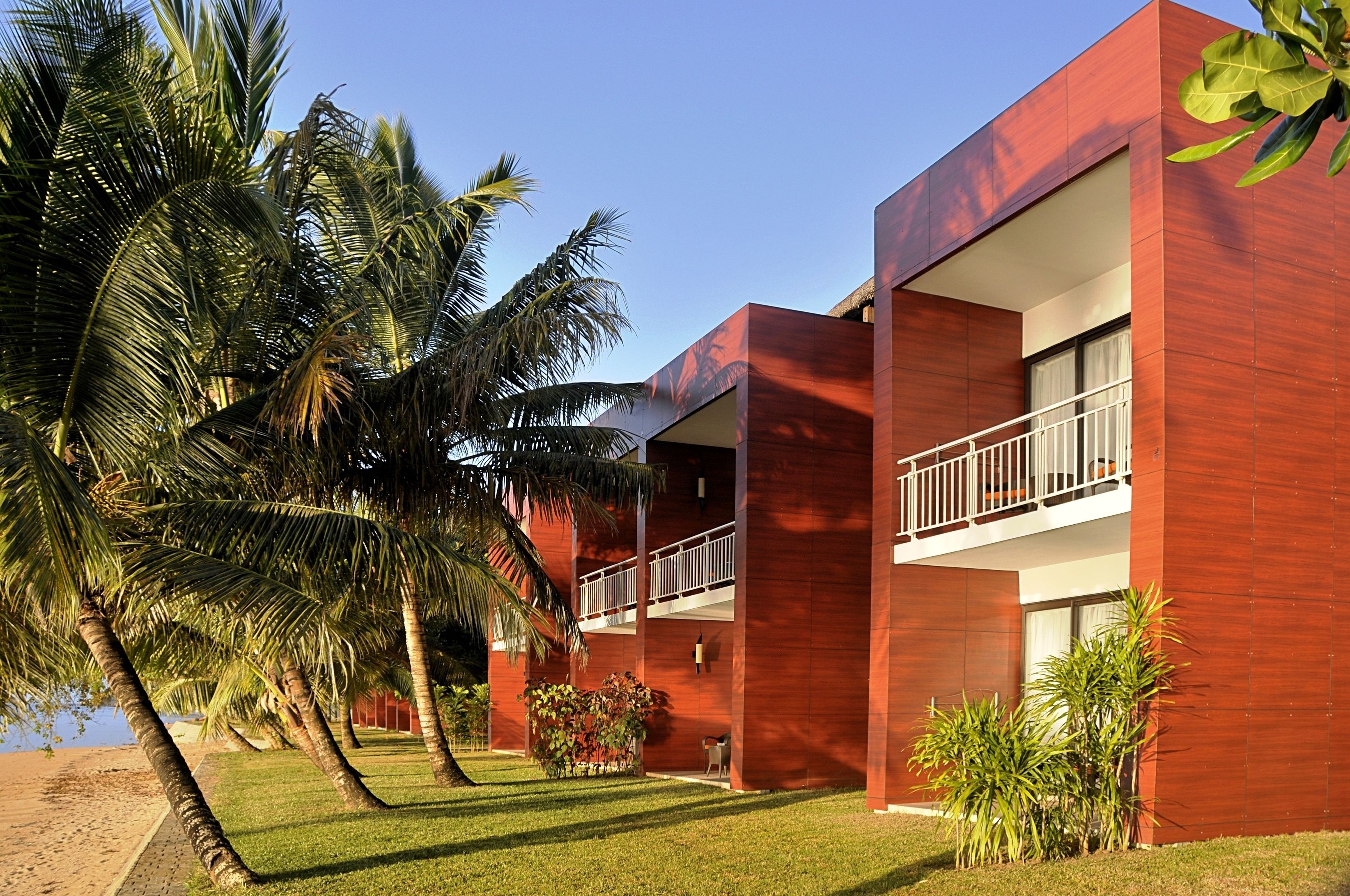 Un edificio moderno con fachada roja, un balcón y palmeras en primer plano.