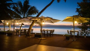 An outdoor dining area with tables and umbrellas, overlooking a body of water at dusk.