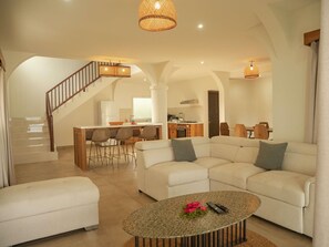 A spacious living area with a white sectional sofa, a round coffee table, and a kitchen in the background. - Mantis Soanambo Hotel and Spa (Nosy Boraha)