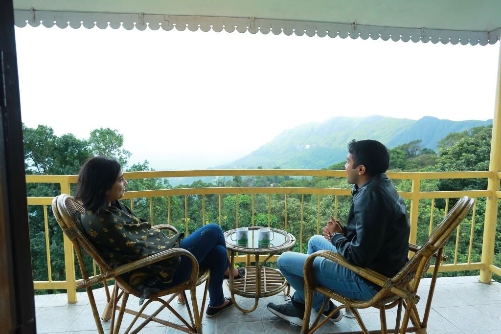 Deux personnes sont assises à une petite table ronde sur un balcon dont la rambarde est jaune, et d’où l’on peut admirer un paysage montagneux.