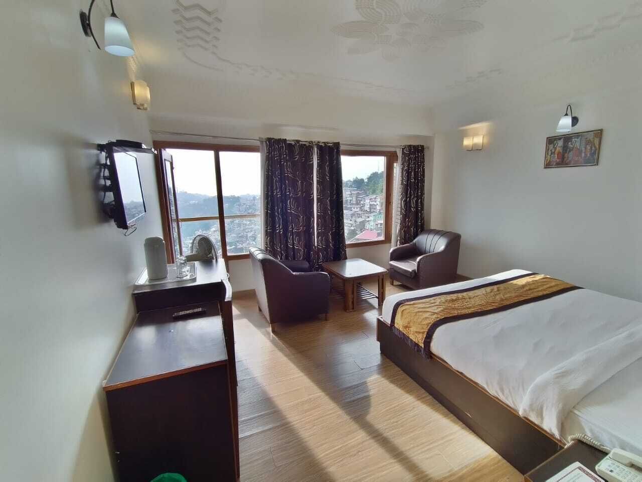 A hotel room with a bed, a desk, a chair, a small table, and a view of the city through large windows.