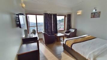 A hotel room with a bed, a desk, a chair, a small table, and a view of the city through large windows.