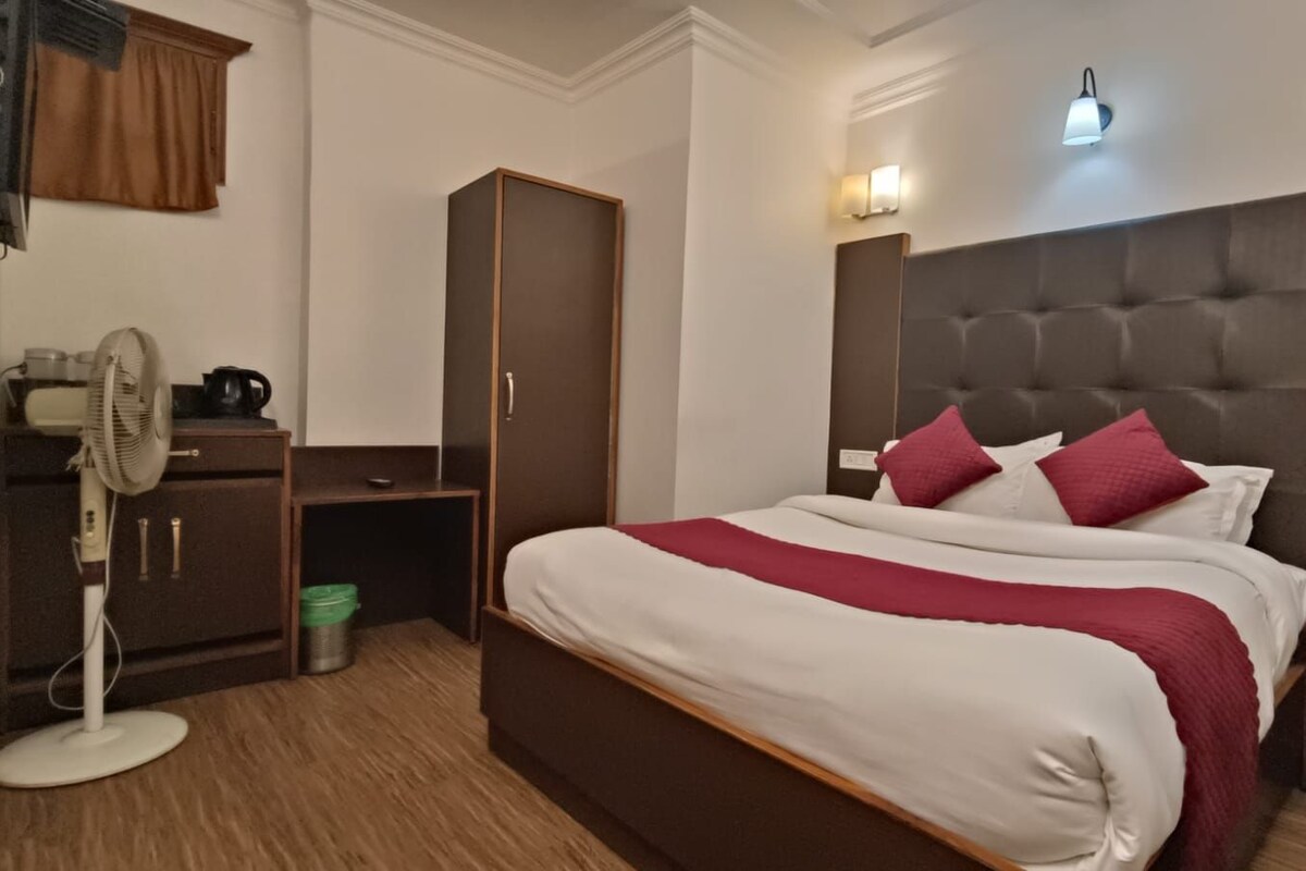 A hotel room with a large bed, a wardrobe, a desk with a fan, and a nightstand with a phone.