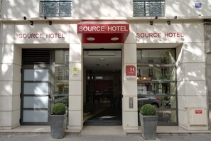 L’entrée de l’hôtel Source, avec sa façade moderne, ses grandes baies vitrées et son entrée centrale.