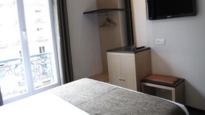 A hotel room with a bed, a small table, a cabinet, a TV, and a window with a view of buildings. - Source Hotel (Paris)
