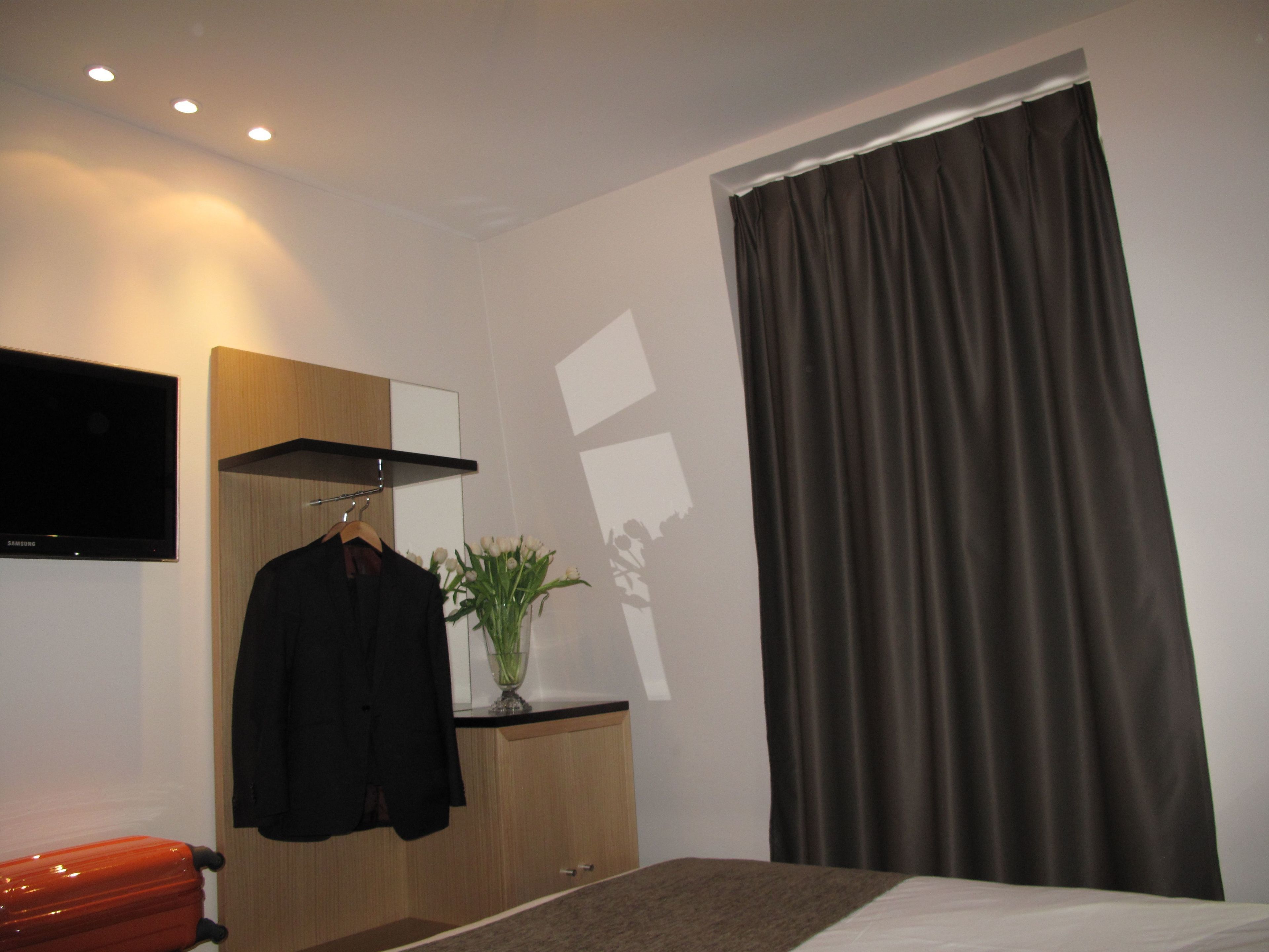A hotel room with a bed, a TV mounted on the wall, a coat hanging on a rack, and a window with curtains.