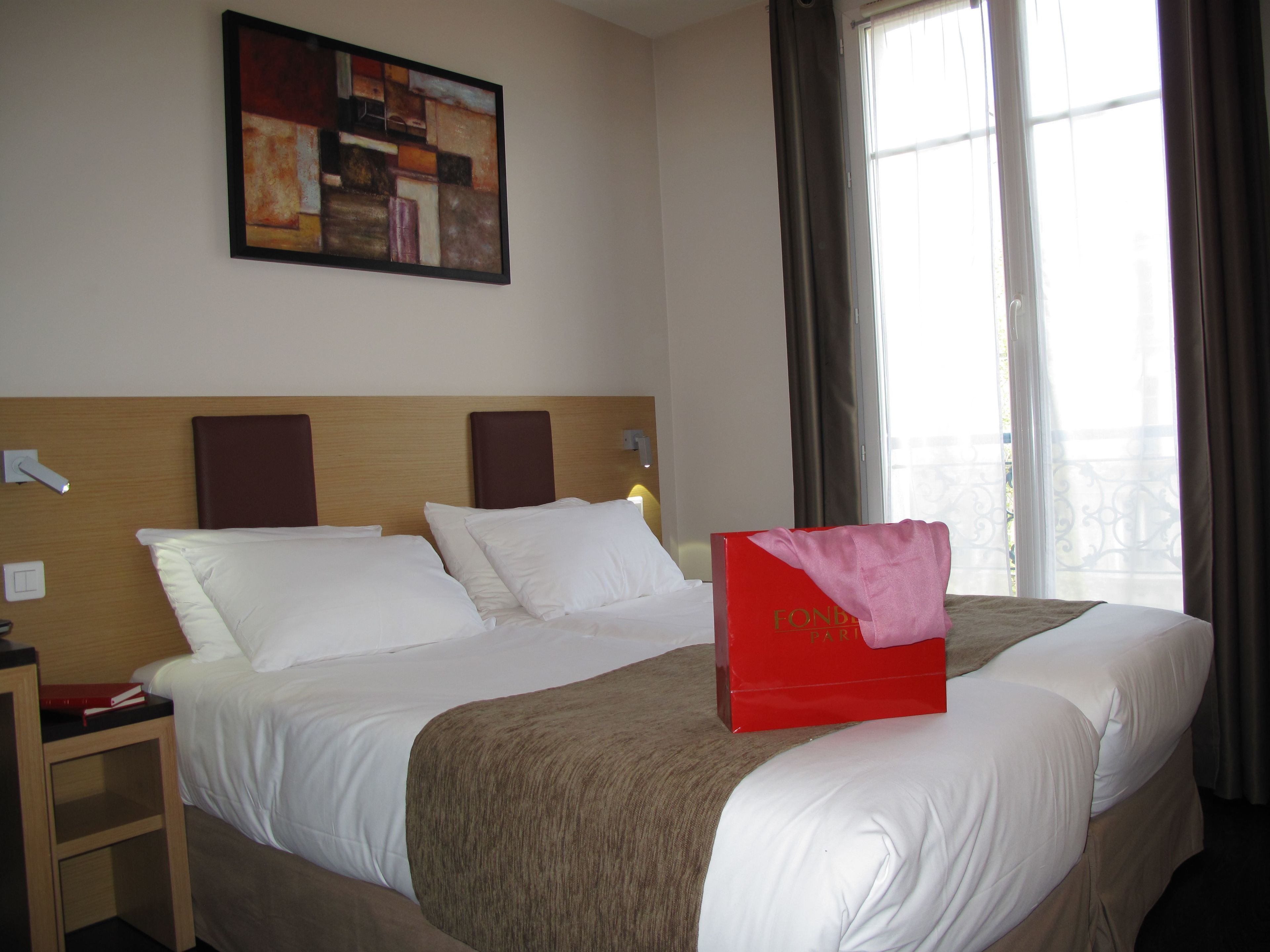 A hotel room with a large bed, a bedside table, a nightstand, a wall-mounted painting, and a window with curtains.