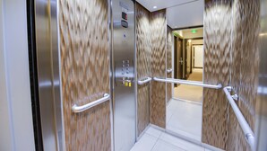 An elevator with metallic walls and a wooden panel design, featuring a control panel and handrails. - Source Hotel (Paris)