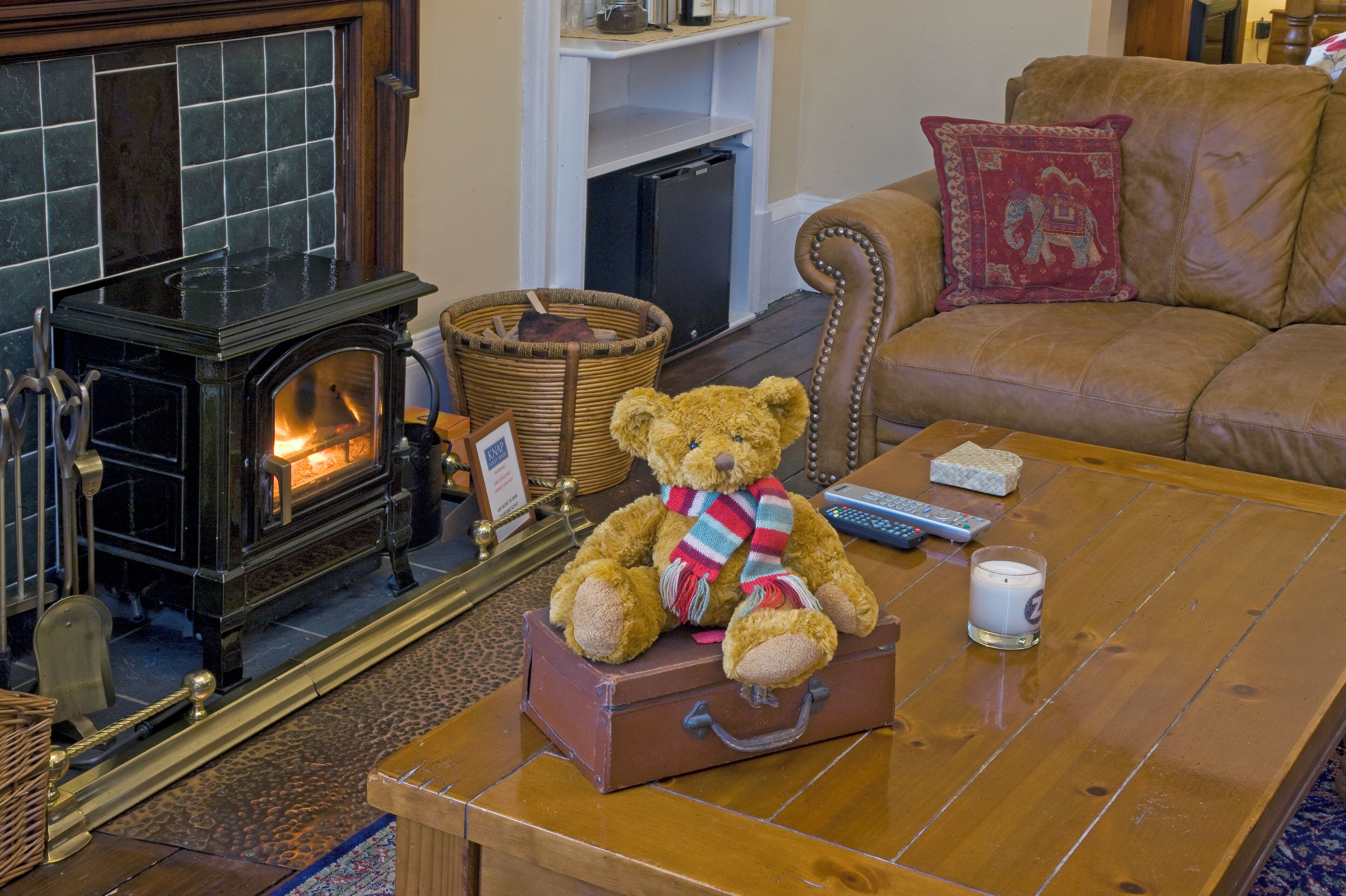 A cozy living room with a fireplace, a brown leather sofa, a coffee table with a teddy bear and a remote control, and a wicker basket.