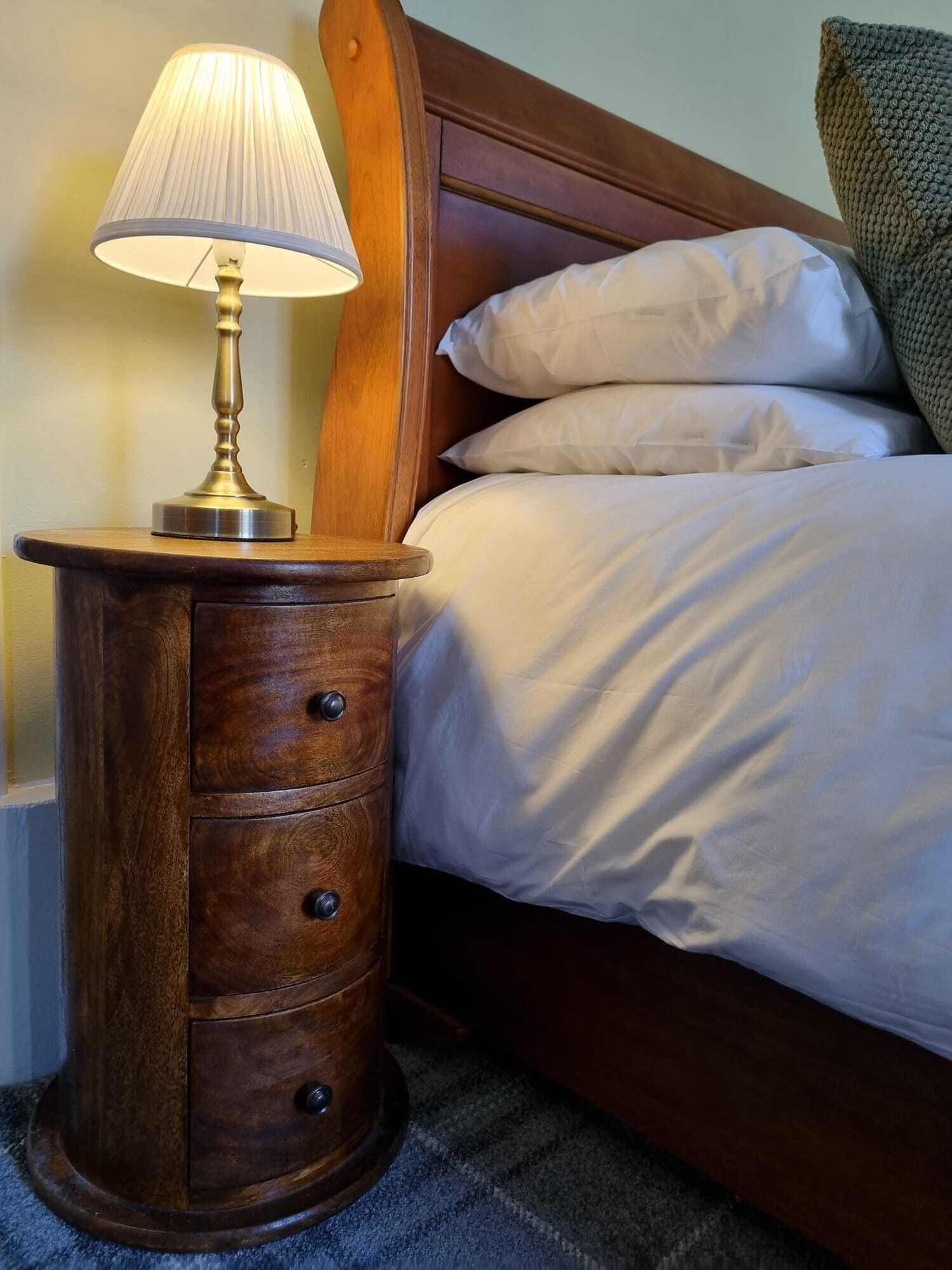 Een houten nachtkastje met een lamp, een bed met witte lakens en een groen gebreid kussen.