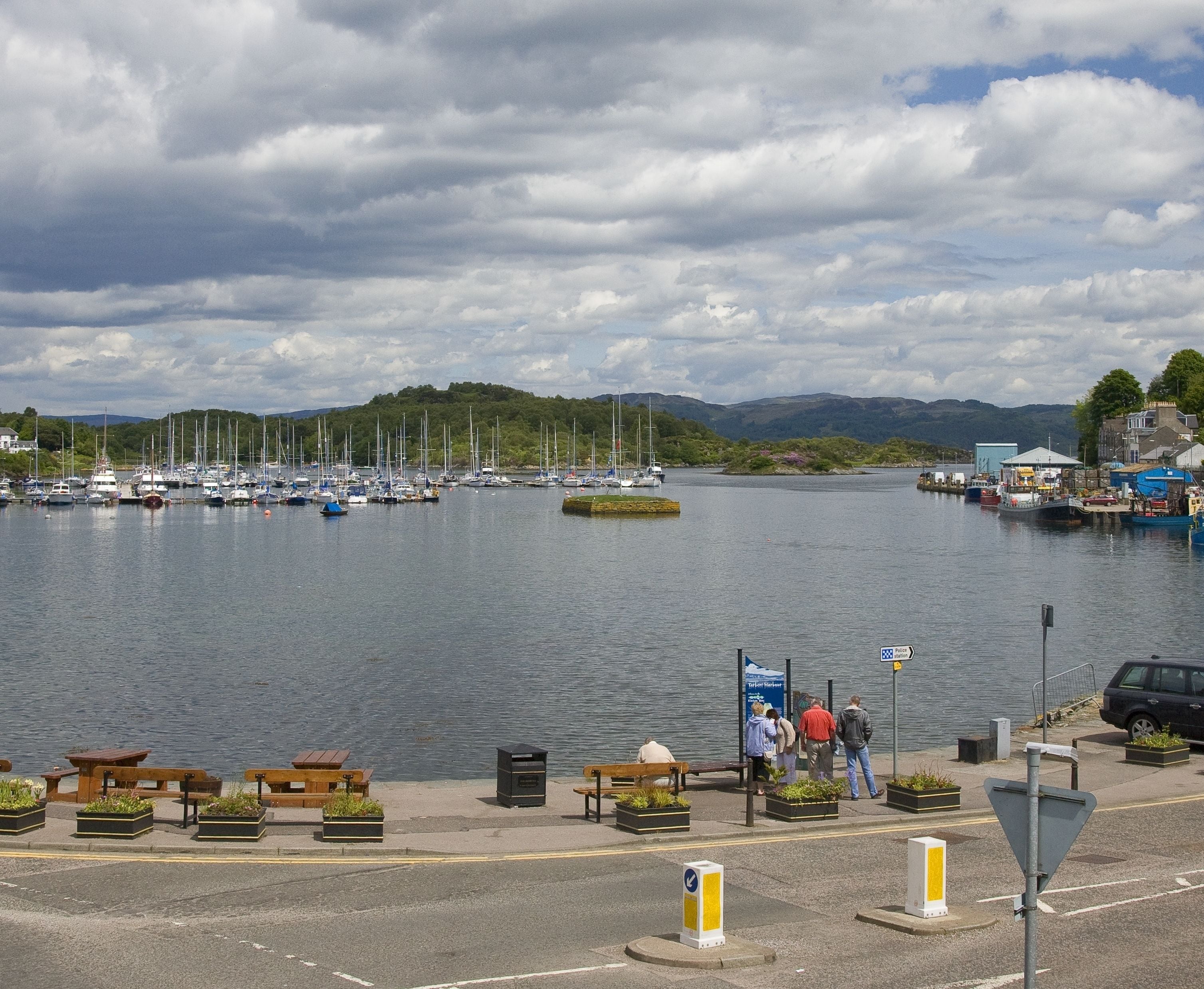 Un port de plaisance avec des bateaux amarrés, une promenade piétonne bordée de bancs et une vue sur des collines en arrière-plan.