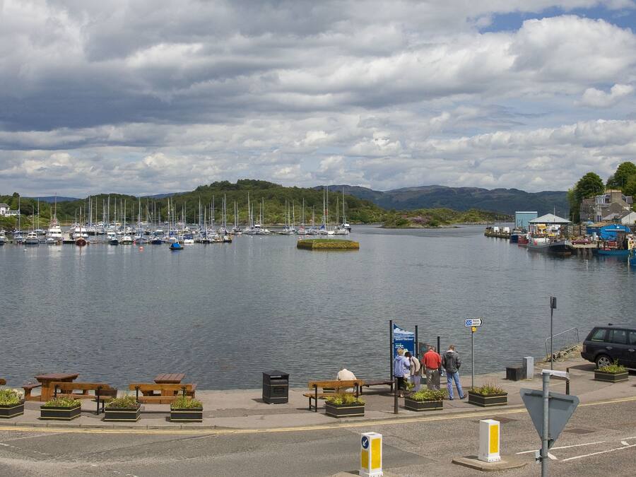 Un port de plaisance avec des bateaux amarrés, une promenade piétonne bordée de bancs et une vue sur des collines en arrière-plan.