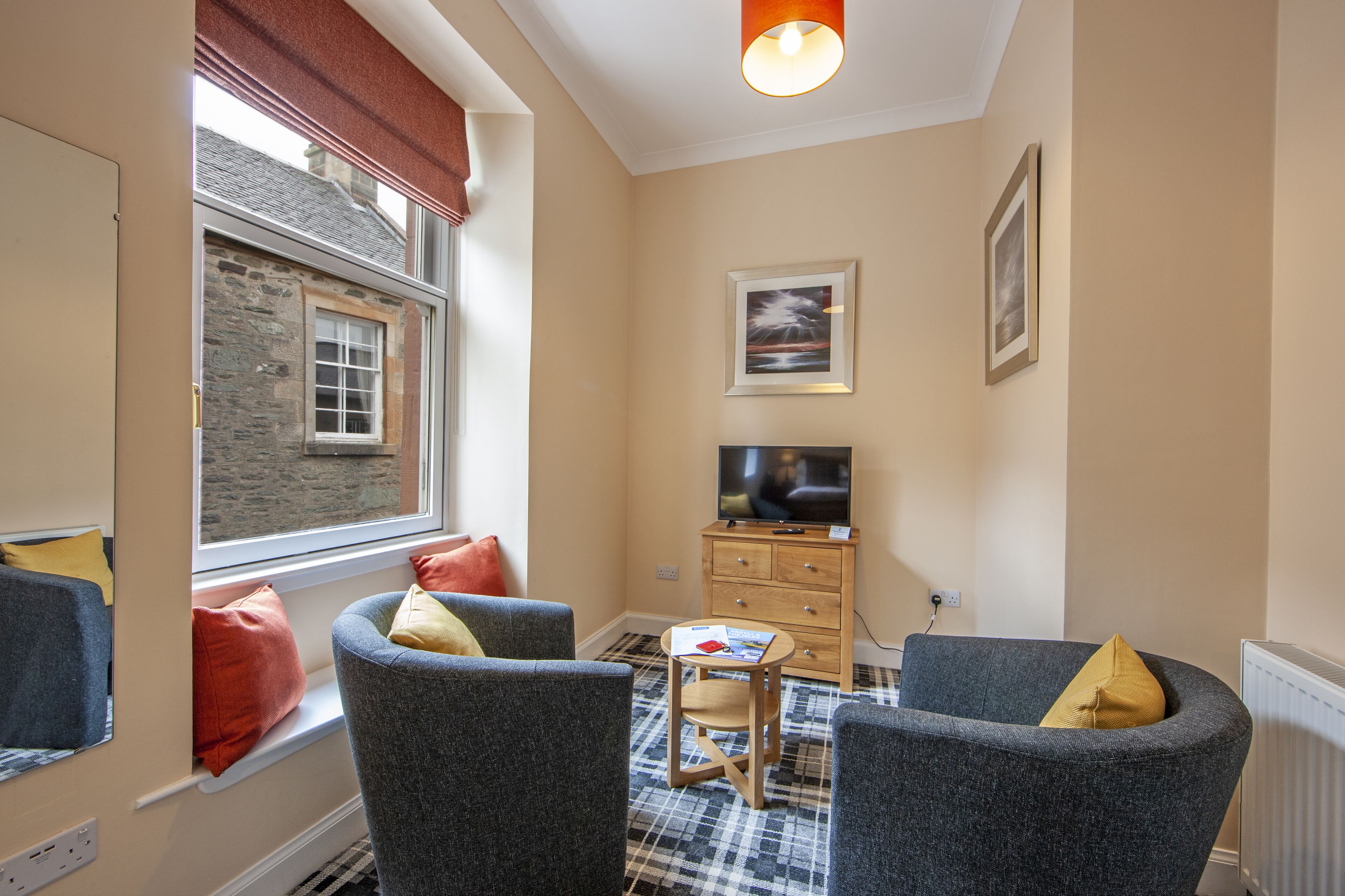 A cozy living room with two grey armchairs, a small wooden table, a television, and a window with a view of a stone building.