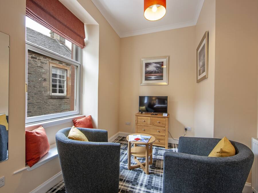 A cozy living room with two grey armchairs, a small wooden table, a television, and a window with a view of a stone building.