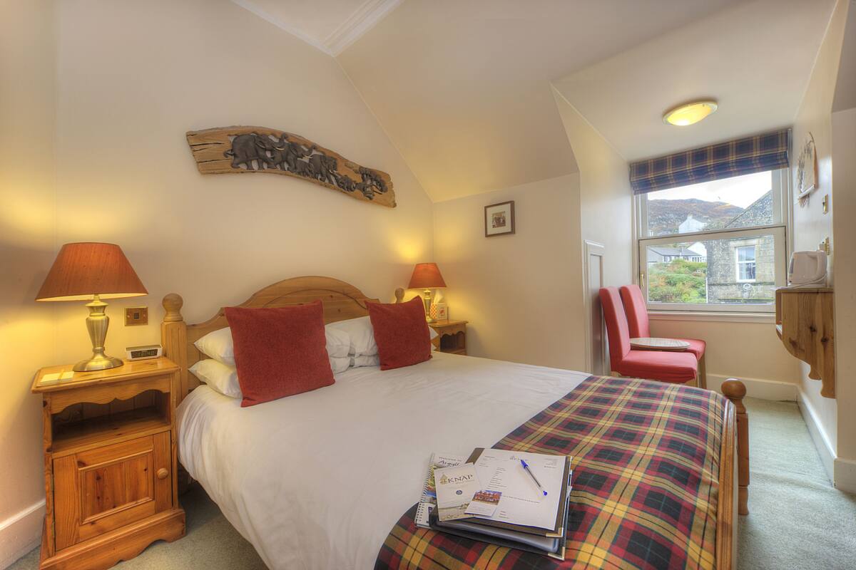 A neatly made bed with a plaid bedspread, two bedside lamps, a wooden headboard, a red chair, and a window with a view of the outside.