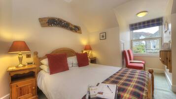 A neatly made bed with a plaid bedspread, two bedside lamps, a wooden headboard, a red chair, and a window with a view of the outside.