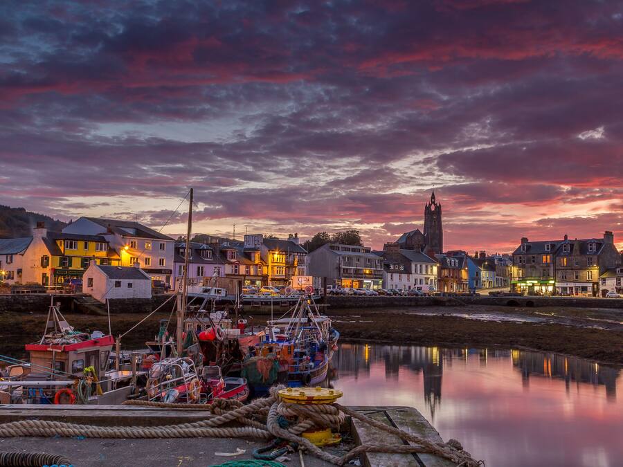 A harbor at dusk with boats docked, a prominent church tower, and buildings with illuminated signs.