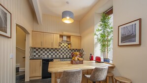 A modern kitchen with wooden cabinets, a checkered backsplash, a central island with bar stools, a hanging light fixture, and a potted plant by the window. - Knap Guest House (Tarbert)