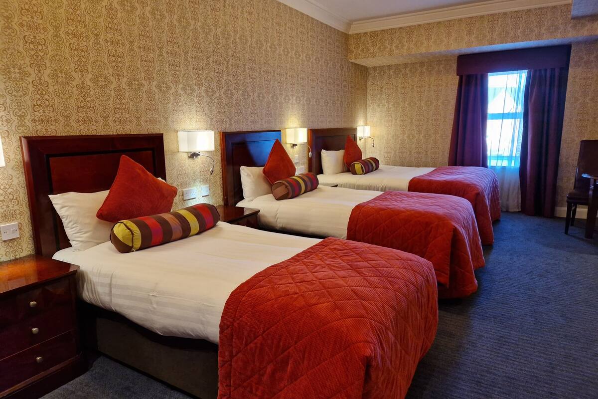A hotel room with three beds, each with red bedding and decorative pillows. The room has patterned wallpaper and a carpeted floor.