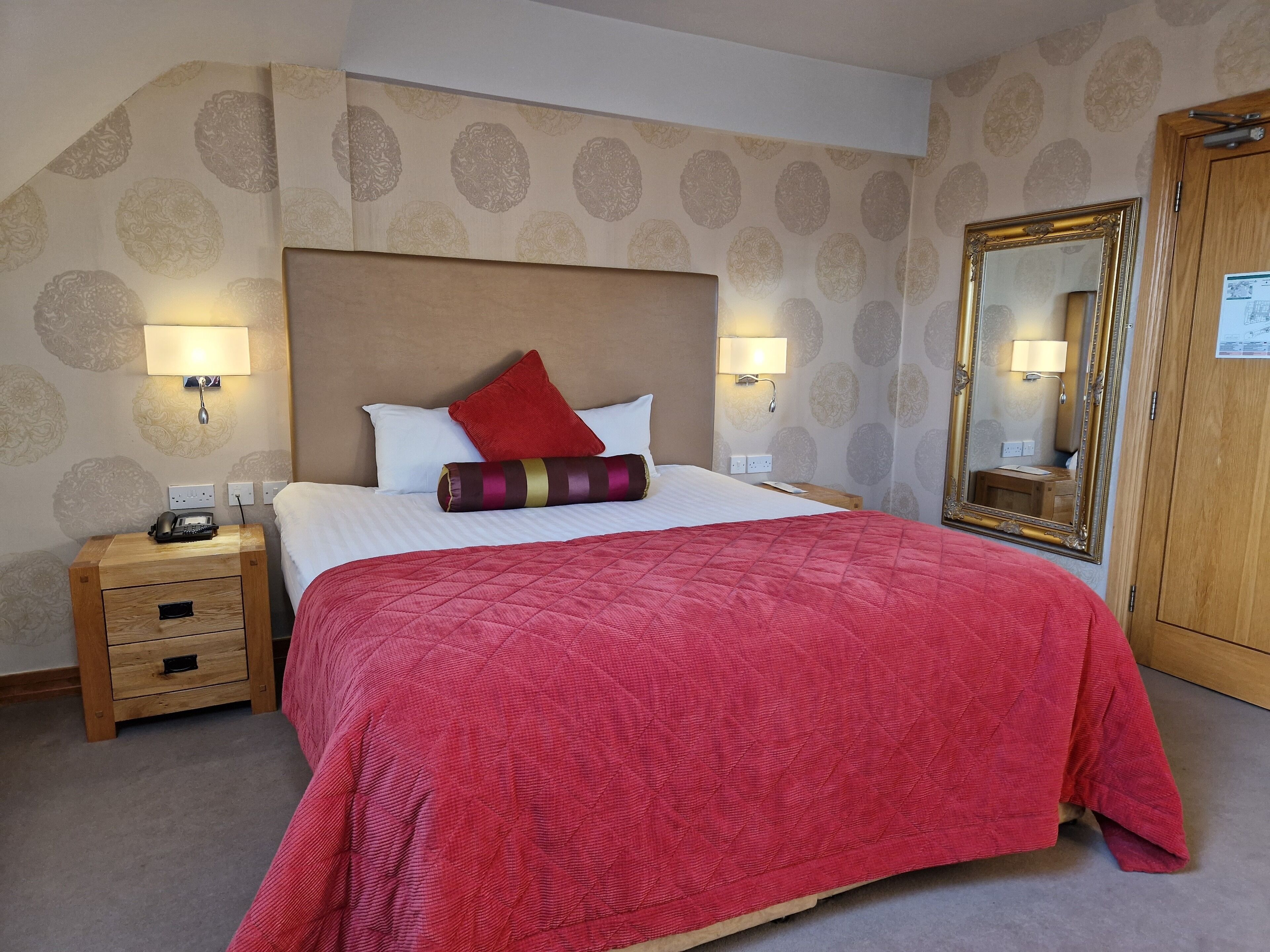 A hotel room with a large bed, a wooden nightstand, a mirror, and a wardrobe.