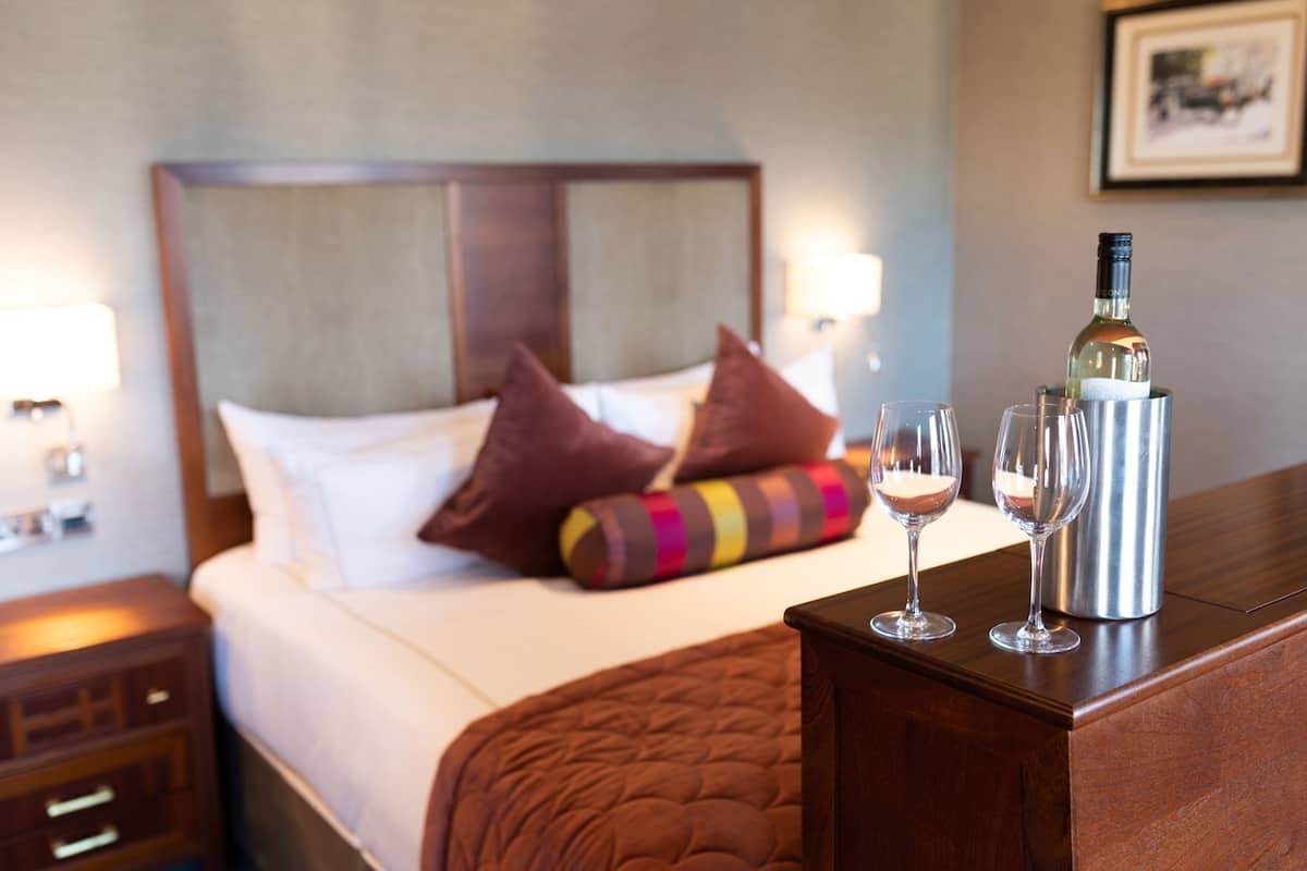 A hotel room with a bed, pillows, a bedside table with a bottle and glasses, and a lamp.