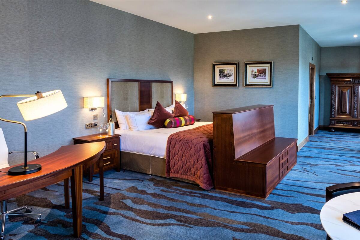 A hotel room with a bed, bedside table, lamp, and a desk. There are framed pictures on the wall and a wooden cabinet in the corner.