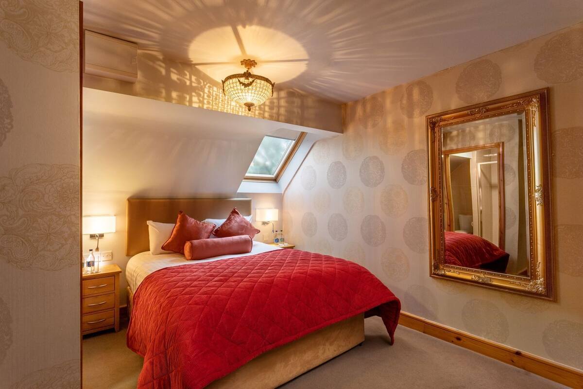 A bedroom with a large bed, a chandelier, a skylight, a mirror, and patterned wallpaper.
