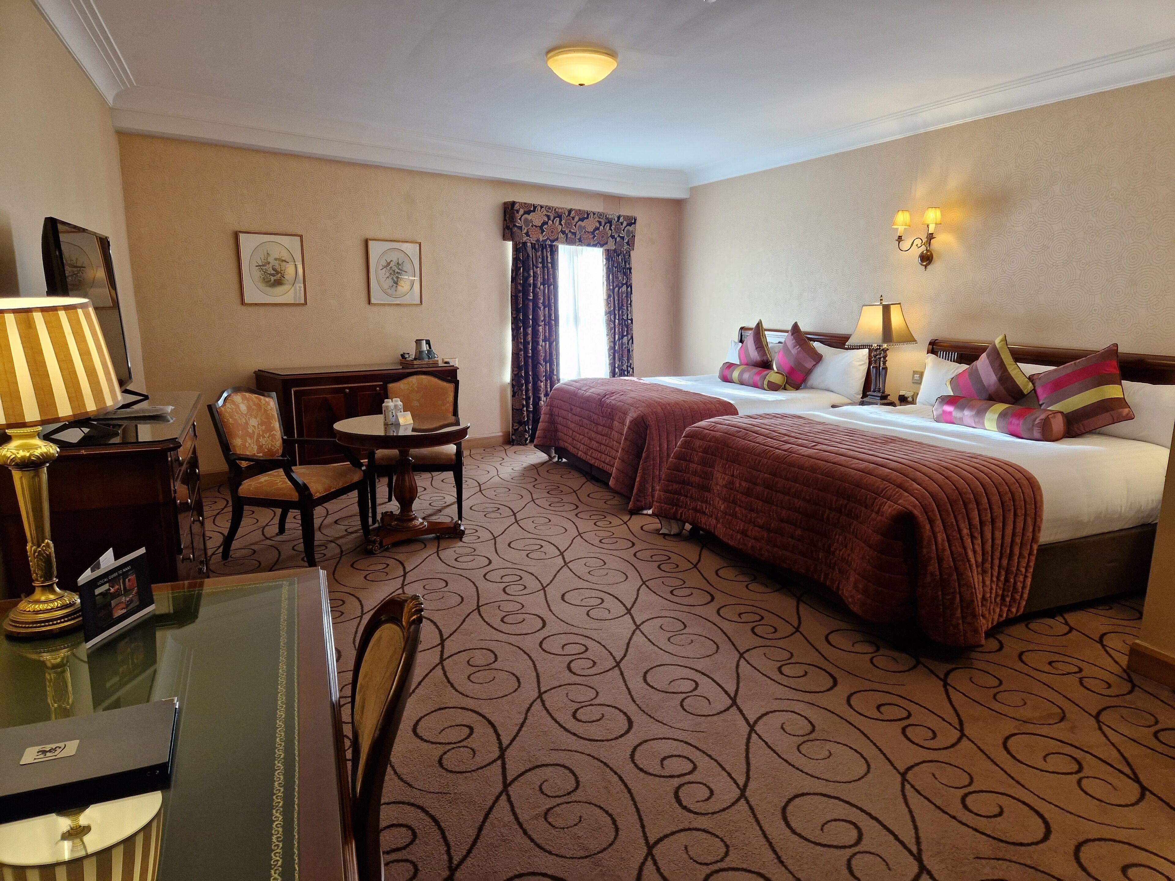 A hotel room with two beds, a desk with a chair, a small table, and a lamp.