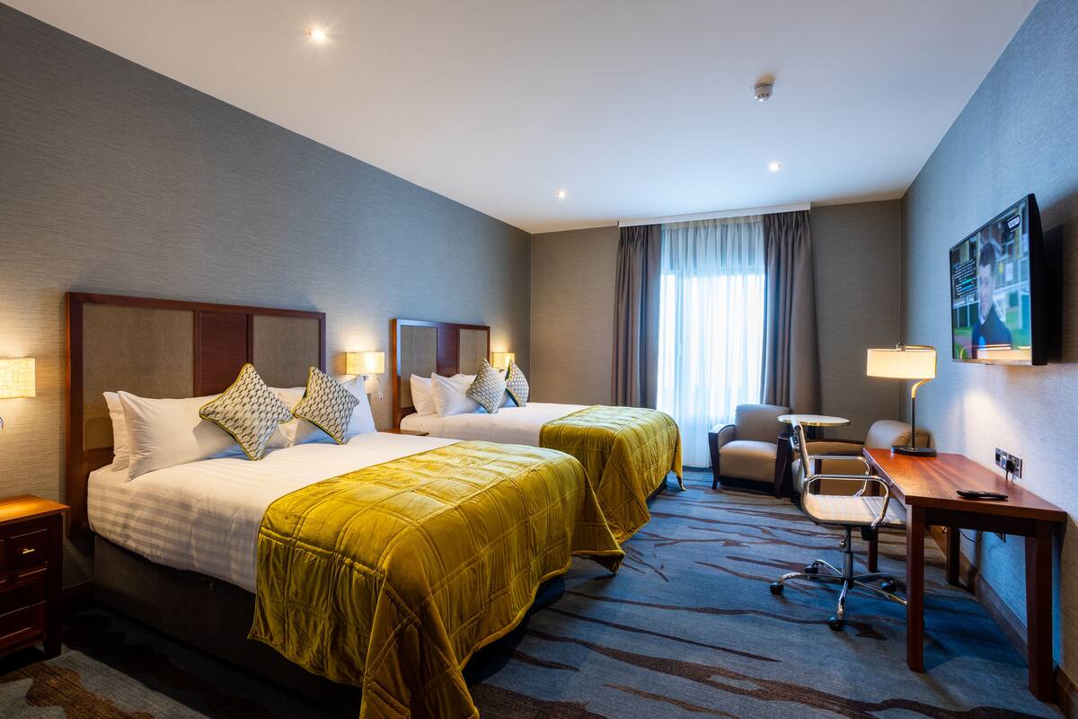A hotel room with two beds, a desk, a chair, a TV, and a window with curtains.