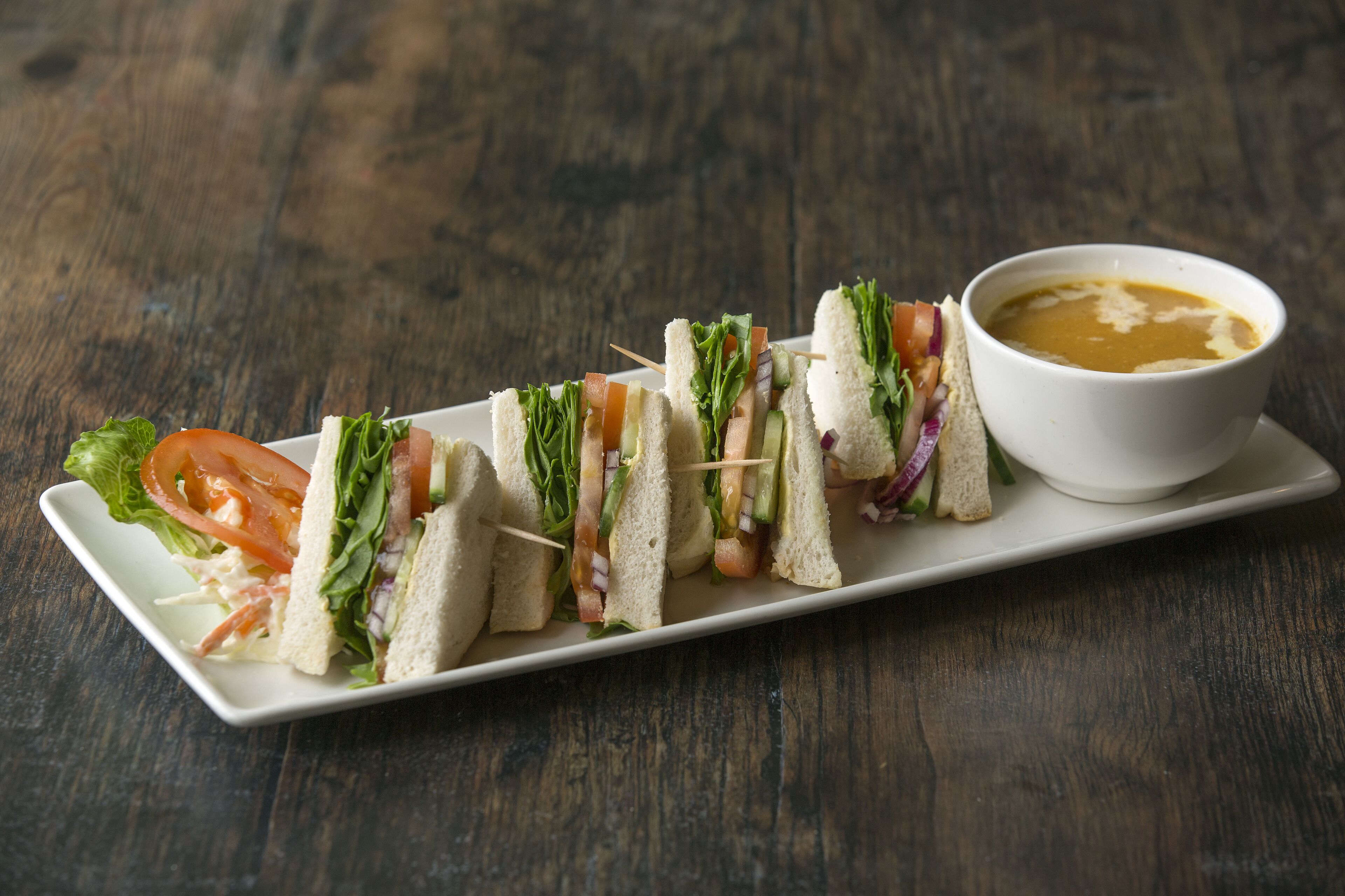 Un plato blanco rectangular con varios pinchos de sándwich y un bol de sopa sobre una mesa de madera.