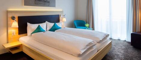 Una habitación de hotel con una cama grande, lámparas de noche, una silla y un ventanal con cortinas.