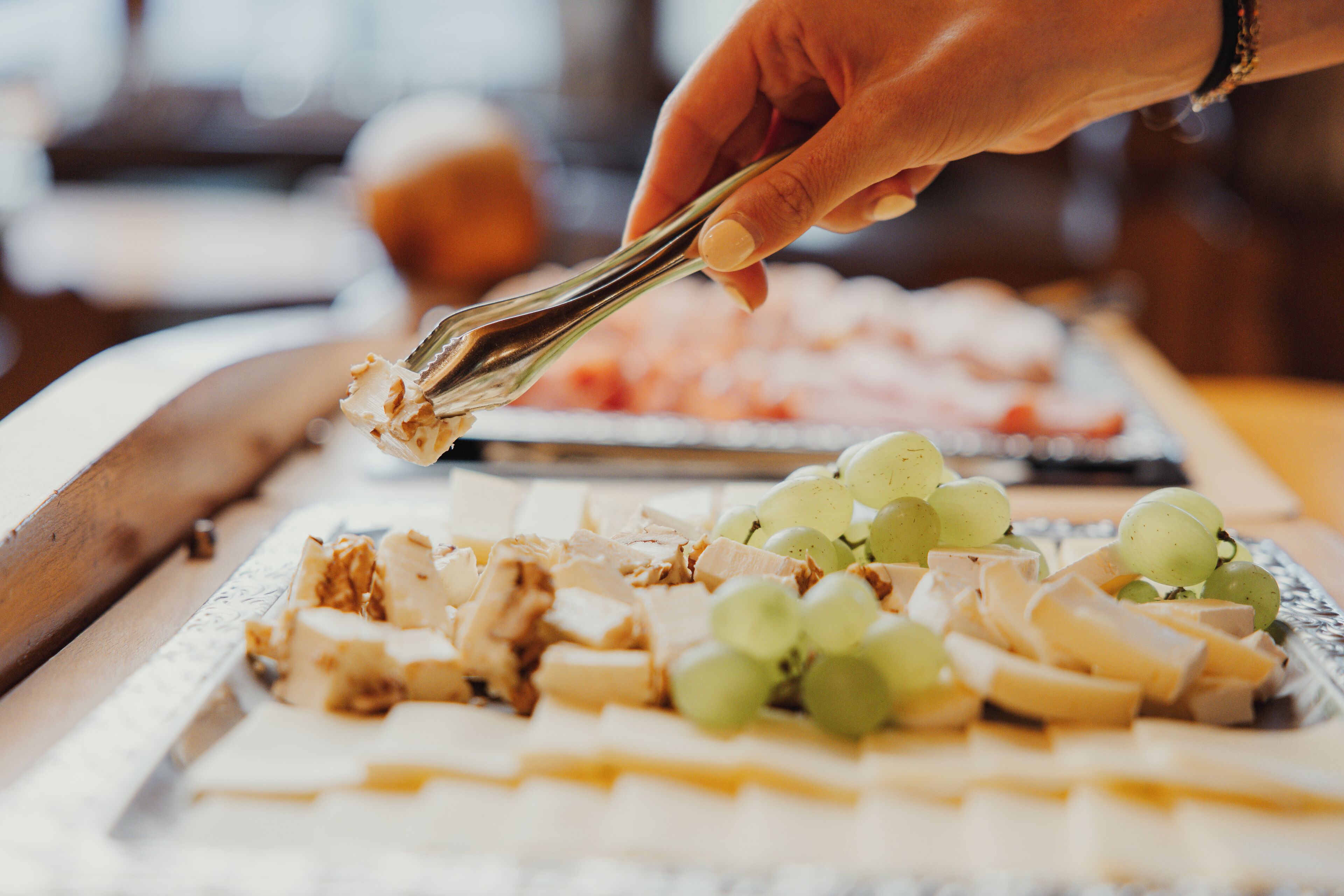 A hand holding tongs over a tray of food, including cheese and grapes.