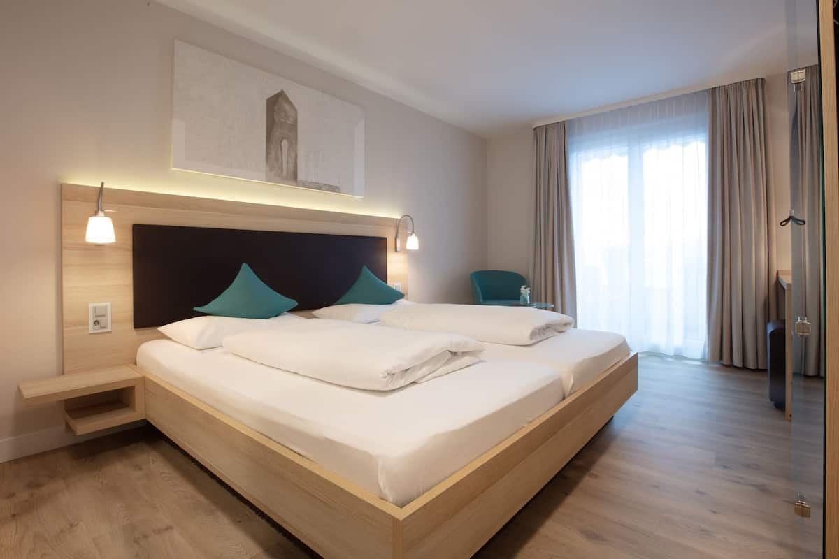 A modern hotel room with a large bed, bedside tables, a wall-mounted artwork, and a view of the outdoors through large windows.