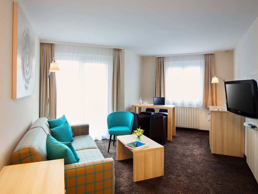 Une chambre d’hôtel avec un canapé, une petite table sur laquelle se trouvent un livre et des fleurs, une télévision et un bureau avec un ordinateur.