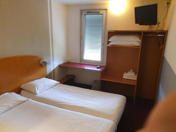 A hotel room with two beds, a small window with blinds, a TV mounted on the wall, and a wooden wardrobe.