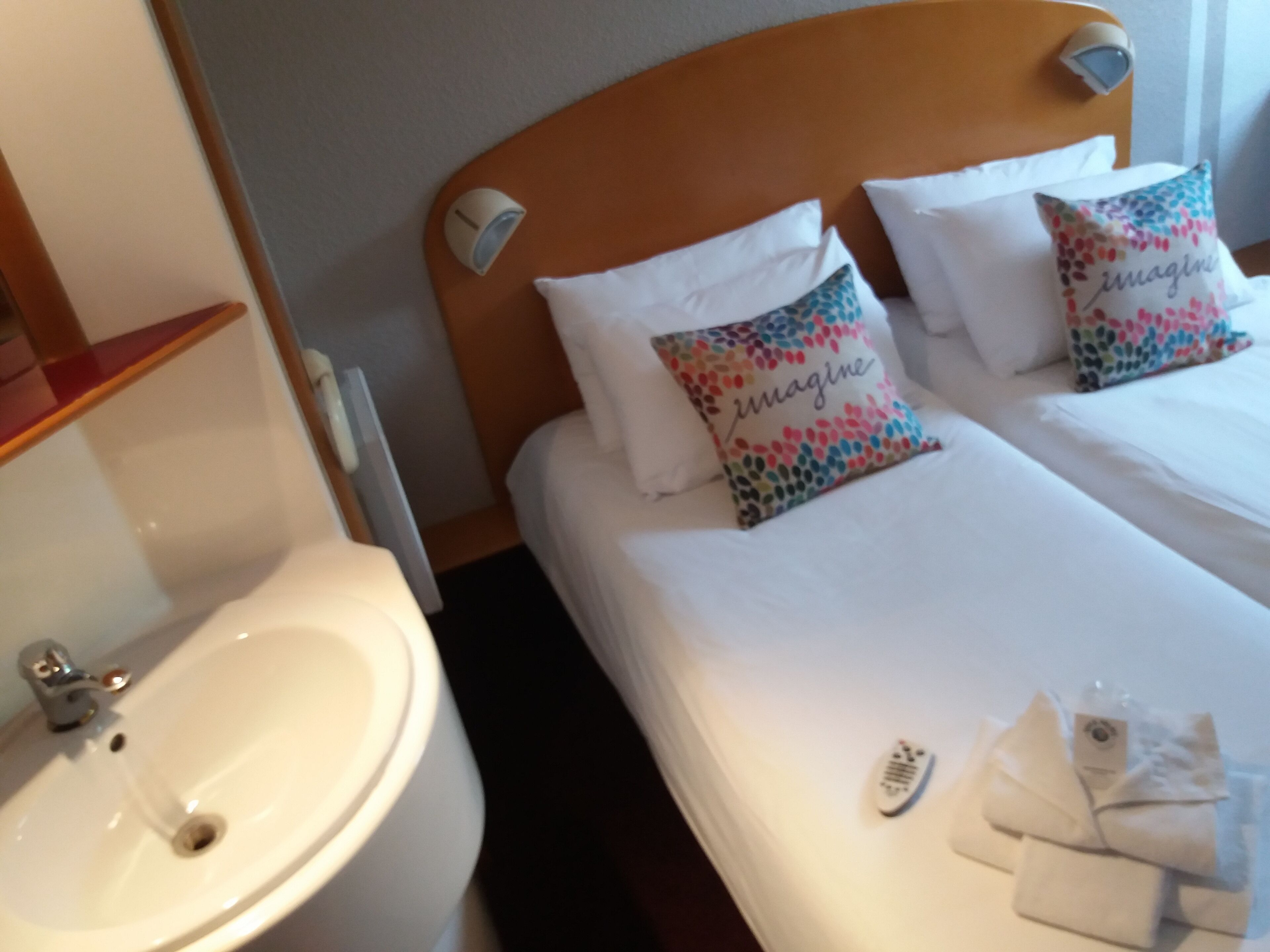 Una habitación de hotel con cabecera de madera, ropa de cama blanca, una mesita de noche con control remoto y un baño con lavabo y espejo.