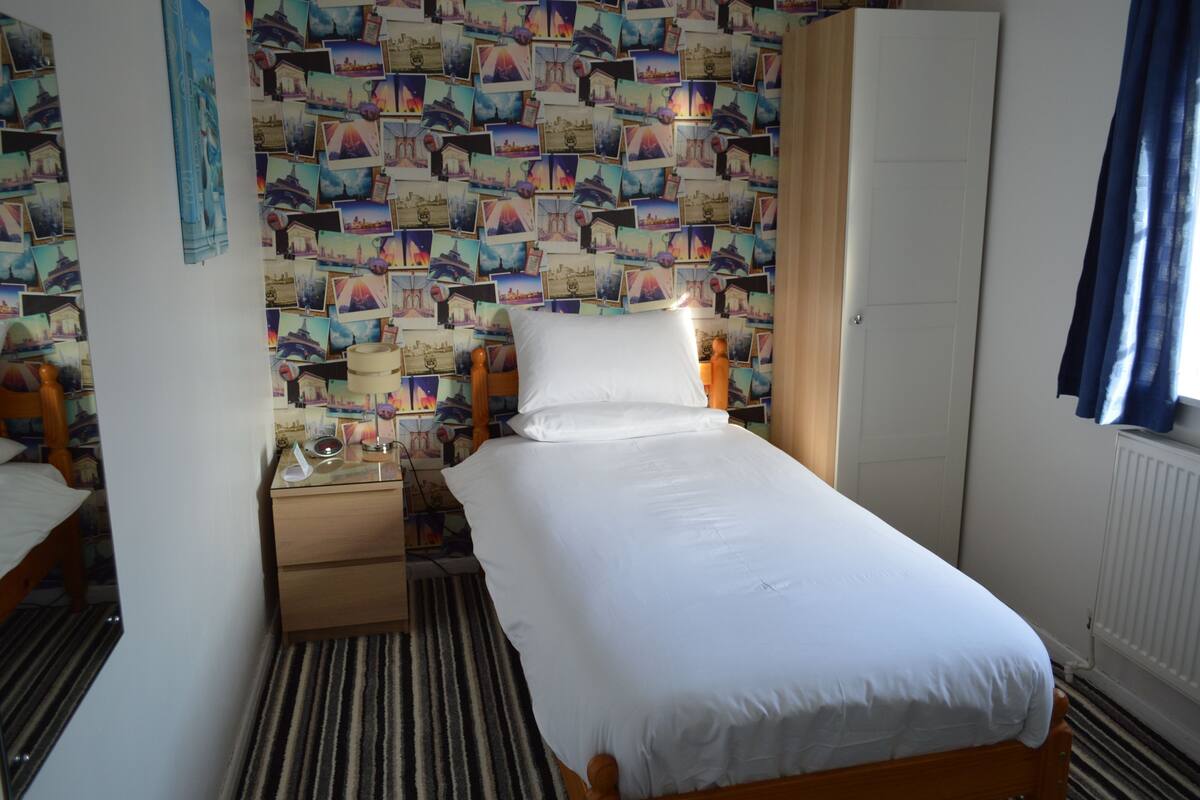A single bed with a white mattress and pillows, a bedside table with a lamp, a wardrobe, and a patterned wall.