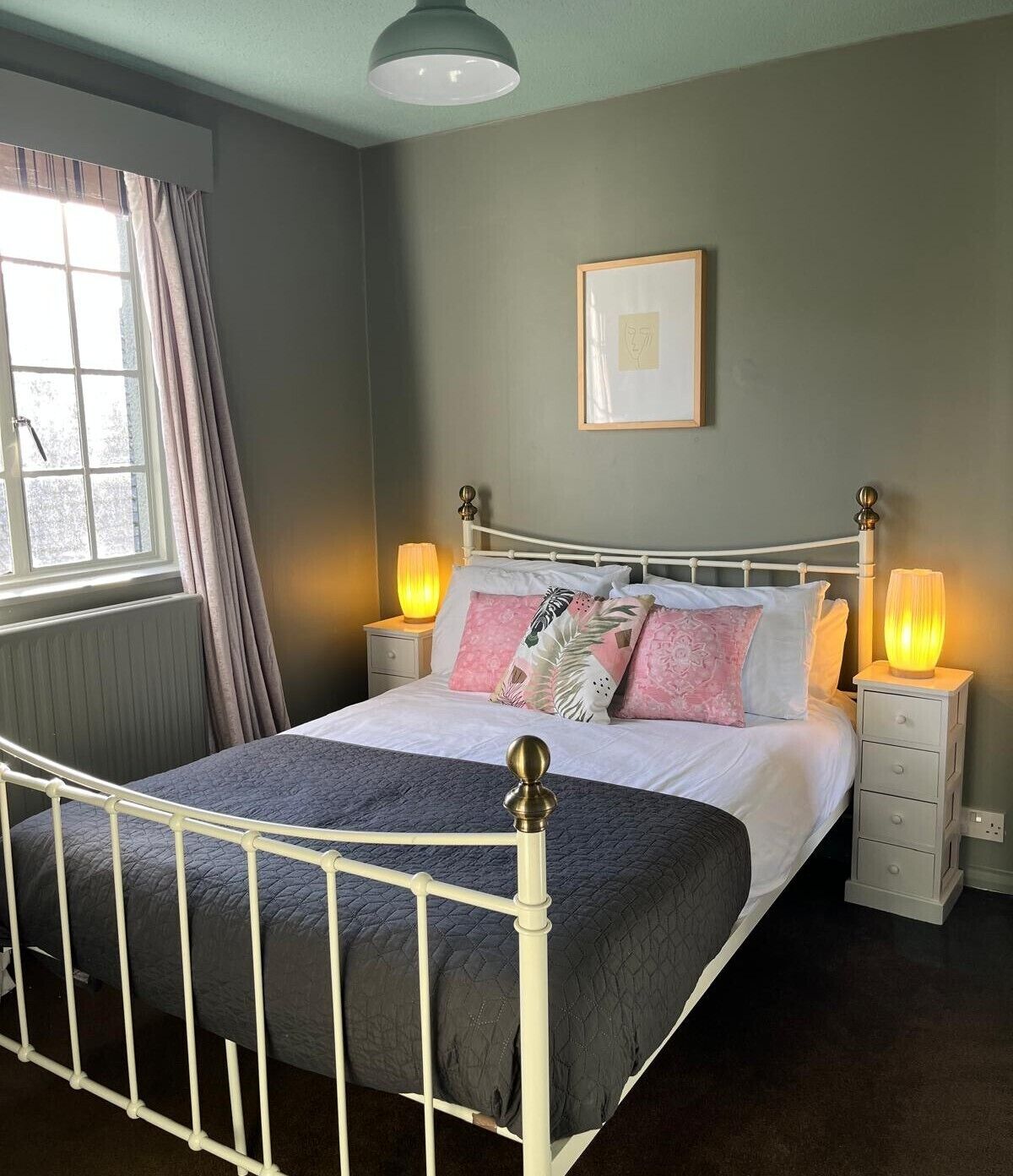 Ein Schlafzimmer mit weißem Metallbettgestell, einer gesteppten grauen Bettdecke, zwei Nachttischlampen, einem Fenster mit Vorhängen und einem gerahmten Bild an der Wand.