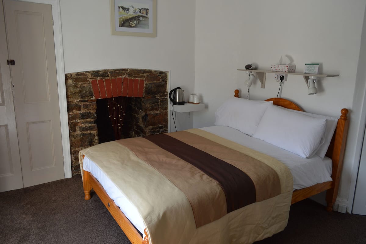 A bedroom with a bed, a brick fireplace, a white door, and a shelf with items.