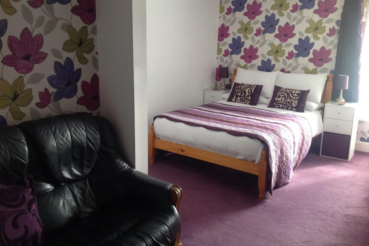 A bedroom with a bed, a nightstand, a lamp, a chair, and floral wallpaper.