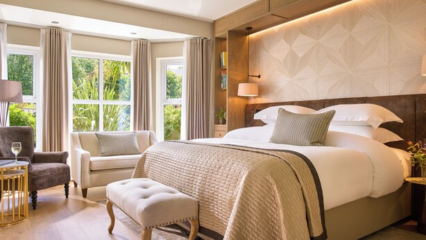 A hotel room with a large bed, a seating area with a chair and ottoman, a bedside table with a lamp, and a view of greenery through the windows. - Brook Lane Hotel (Kenmare)